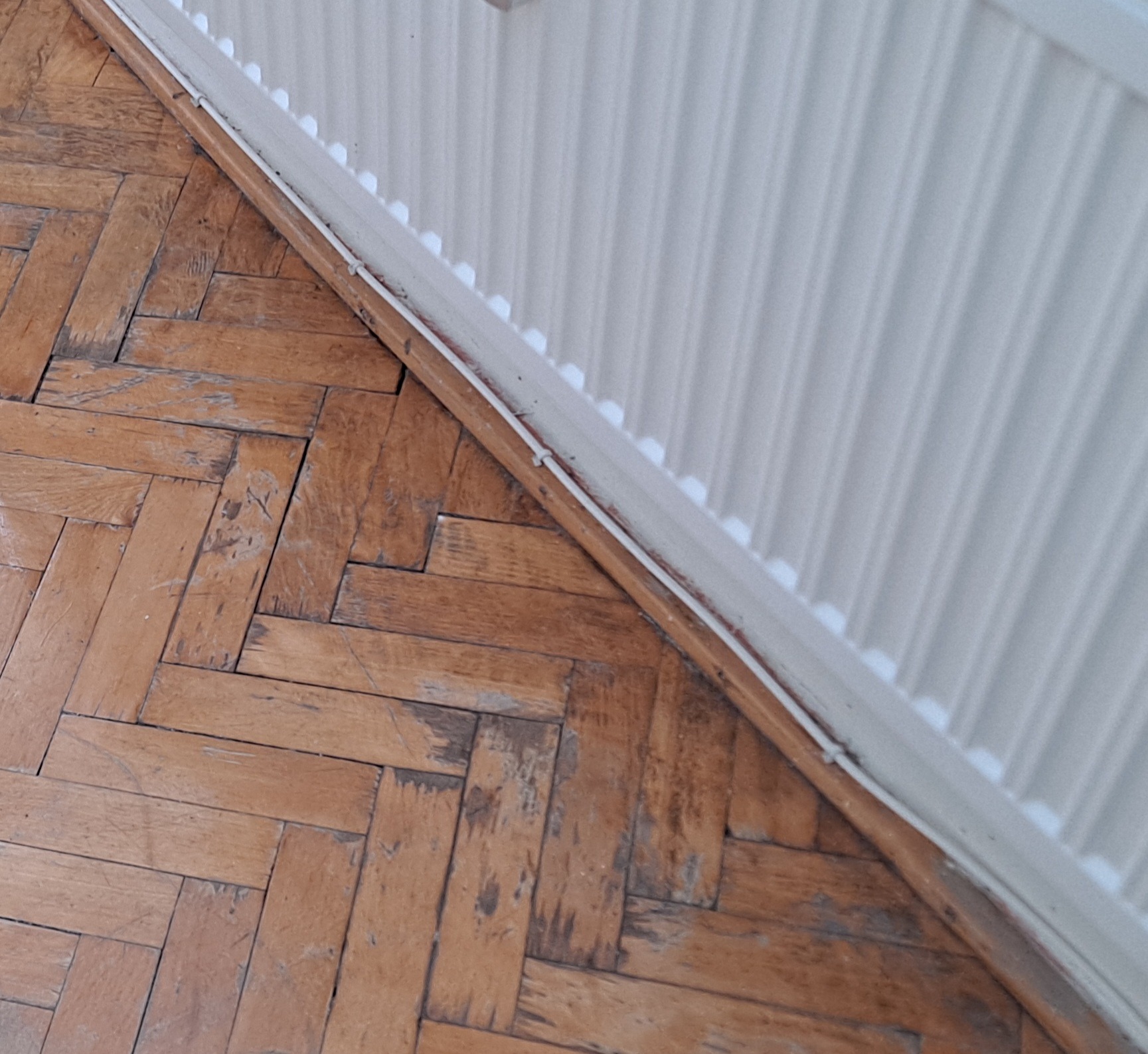 Stara, zniszczona podłoga parkietowa ułożona w jodełkę, z widocznymi śladami użytkowania i ubytkami, przylegająca do białego grzejnika żeberkowego.