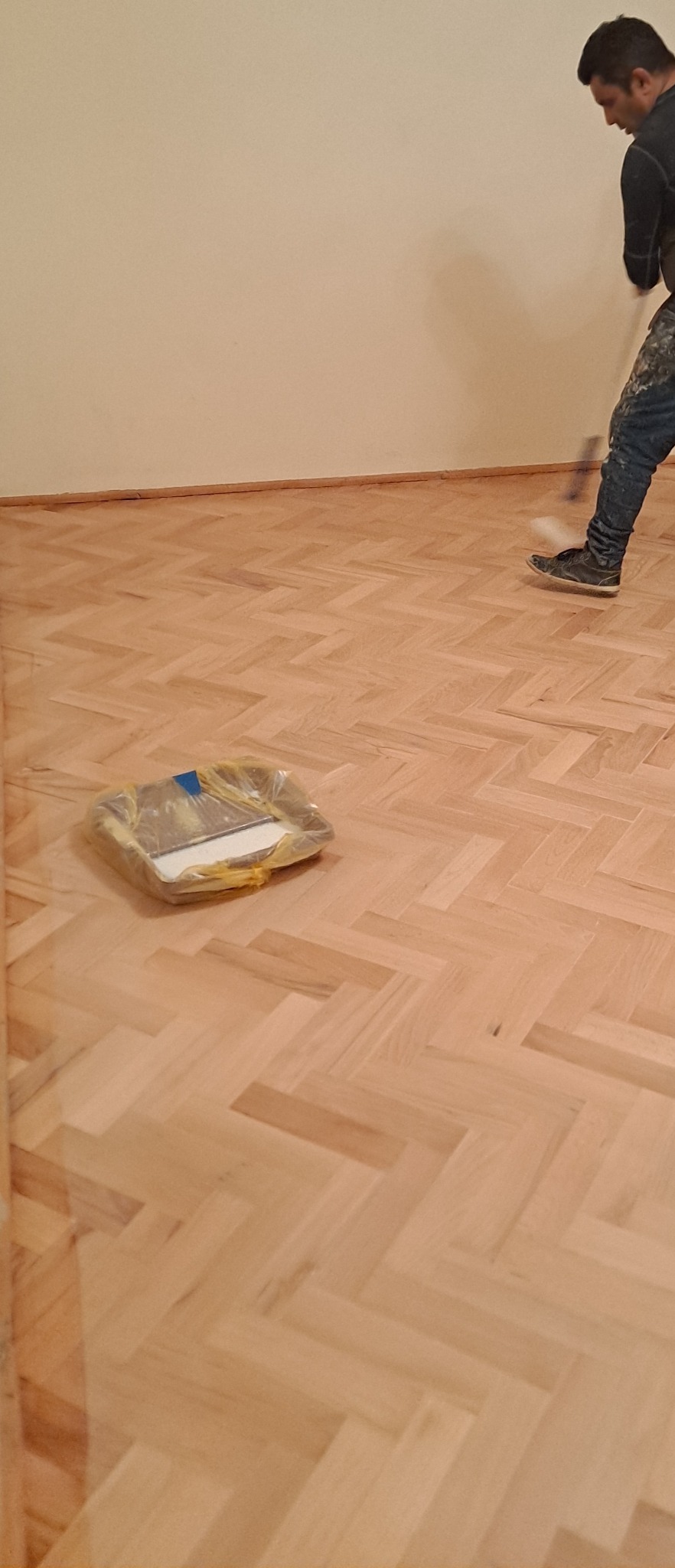 Parkiet ułożony w jodełkę, świeżo polakierowany, z widocznym pracownikiem malującym wałkiem przy ścianie i kuwetą z lakierem w folii.
