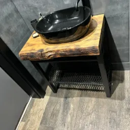 Czarna umywalka nablatowa na blacie z naturalnego drewna z metalowym stelażem w łazience z szarą płytką.