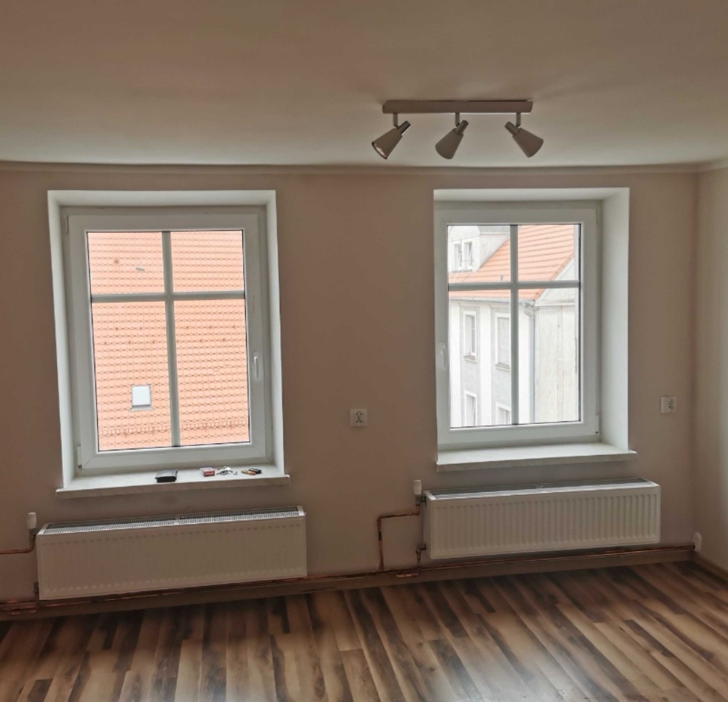Puste pomieszczenie z dwoma oknami z białymi ramami, grzejnikami pod oknami, drewnianą podłogą i trzypunktową lampą sufitową.