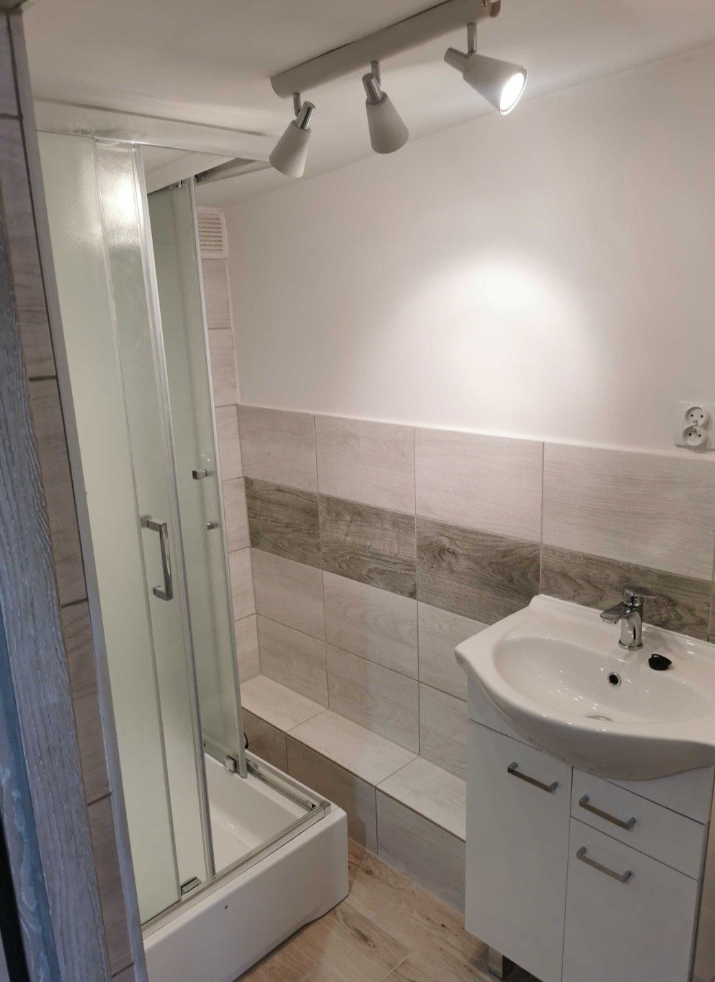 Nowoczesna łazienka po odświeżeniu: kabina prysznicowa z przesuwnymi drzwiami, umywalka z szafką w kolorze białym, płytki ścienne z motywem drewna i oświetlenie punktowe na suficie.