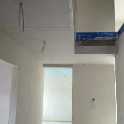 Wnętrze w trakcie prac wykończeniowych: widoczne profile metalowe, płyty na suficie, instalacje elektryczne i ściany przygotowane do malowania, zabezpieczone taśmą malarską.