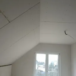 Montaż płyt gipsowo-kartonowych na poddaszu ze skosami, widoczne łączenia płyt i przewód elektryczny wystający z sufitu.