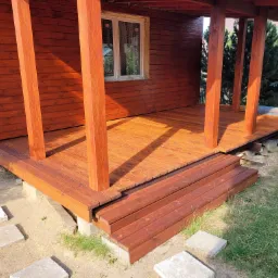 Zadaszony taras drewniany z trzema stopniami, pomalowany na brązowo, z widocznymi słupami podtrzymującymi dach i ścieżką z betonowych płyt.