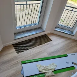 Jasne panele podłogowe w Białymstoku, z widoczną kratką wentylacyjną pod oknem i fragmentem opakowania paneli z napisem 'POSTAW NA WZORNICTWO'.