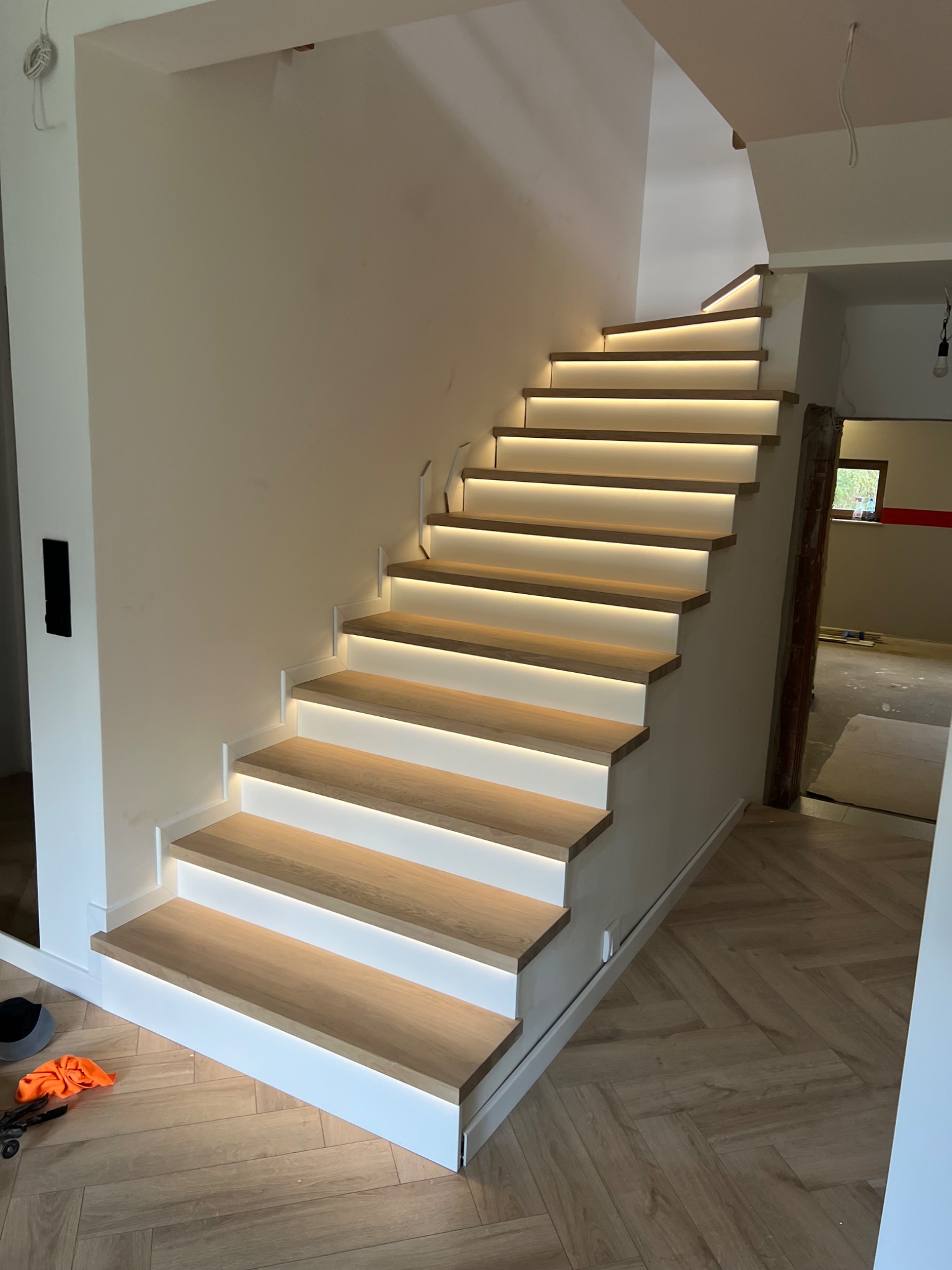 Nowoczesne schody drewniane z subtelnym oświetleniem LED, widok z dołu, jasne wnętrze, parkiet w jodełkę na podłodze, minimalistyczny design.