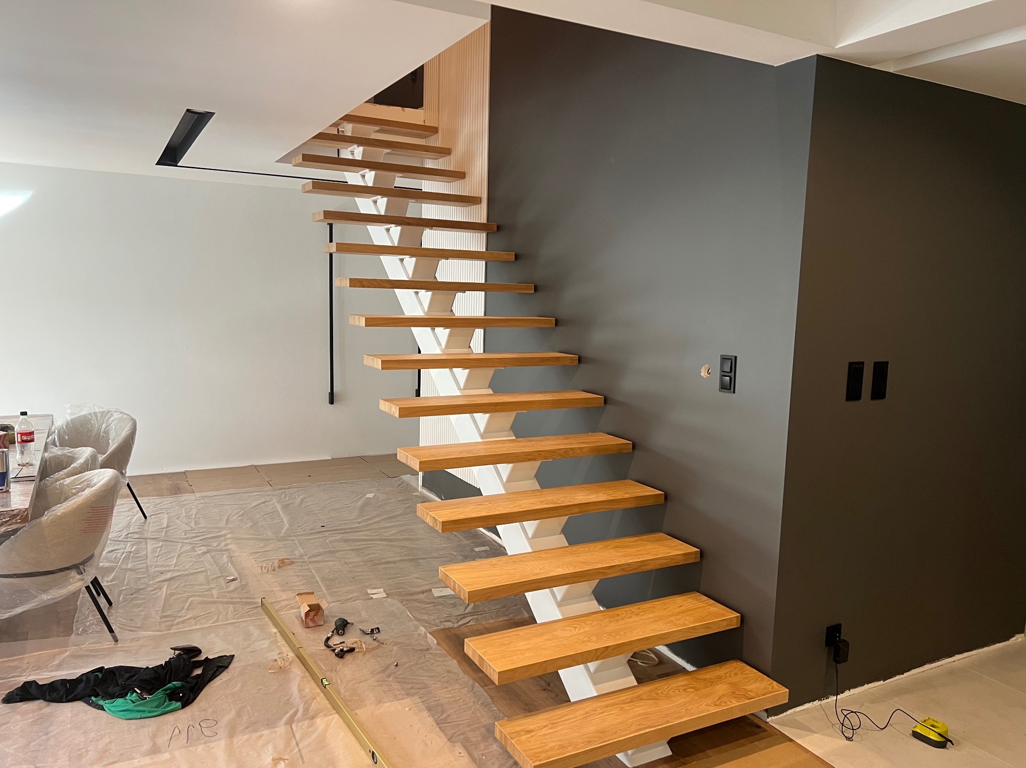 Nowoczesne schody drewniane z białymi wspornikami, widoczne narzędzia i folia ochronna na podłodze, sugerujące etap montażu w jasnym wnętrzu.