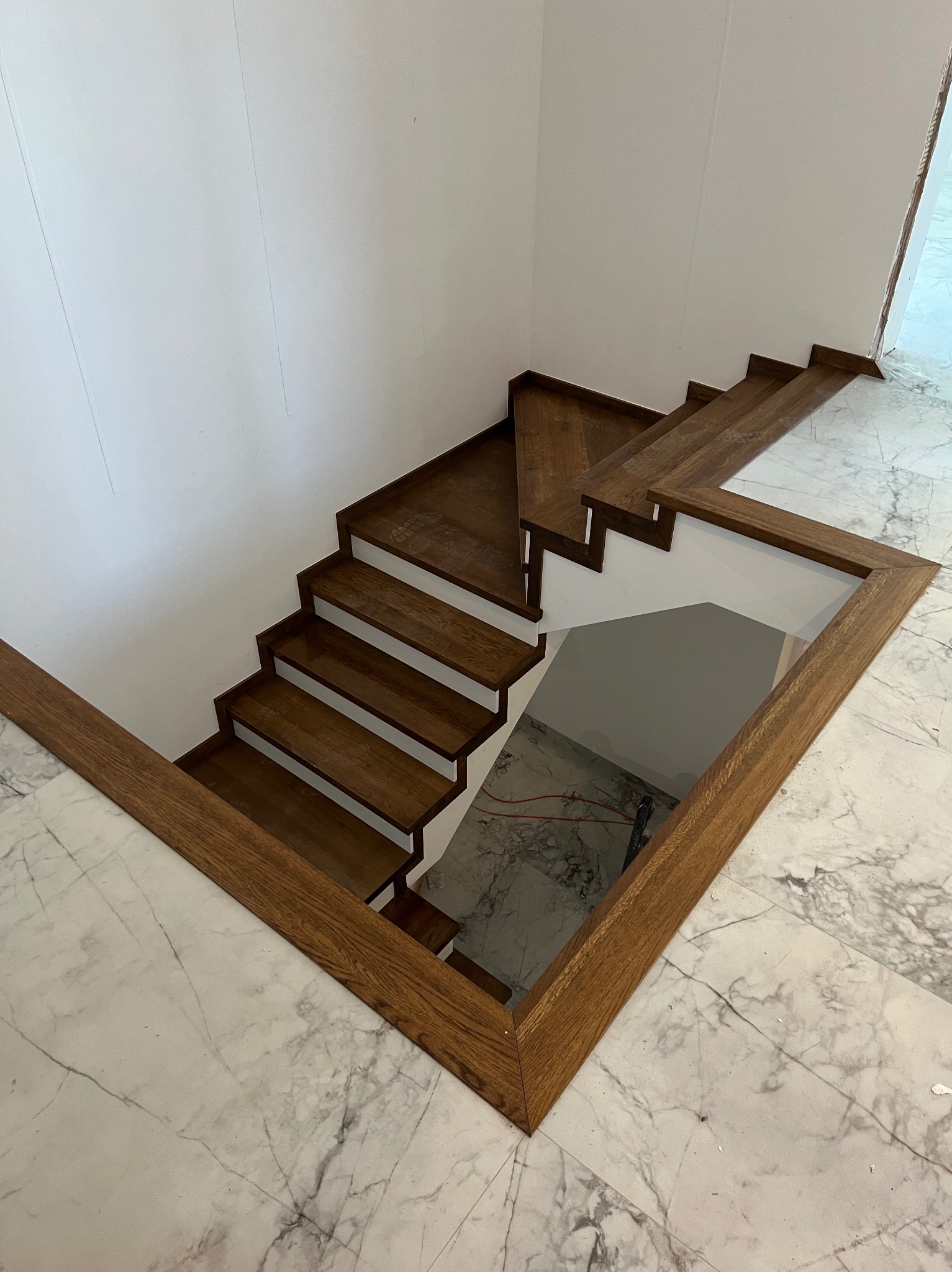 Nowoczesne schody drewniane z ciemnego drewna, widok z góry na marmurową podłogę. Minimalistyczny design, jasne ściany, kątowe ujęcie.