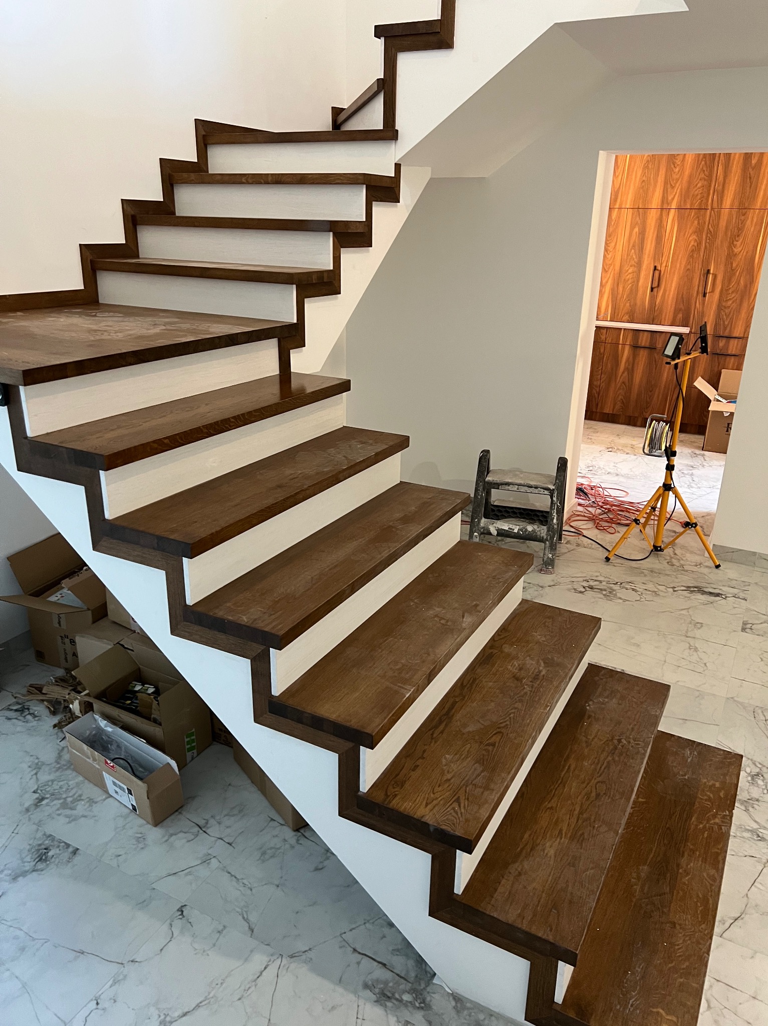Nowoczesne schody z ciemnego drewna i białymi podstopnicami, widoczne z dołu, z narzędziami i oświetleniem w tle, sugerujące etap montażu.
