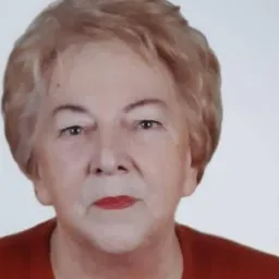 Portret starszej kobiety z krótkimi, jasnymi włosami, w czerwonej bluzce, z delikatnym uśmiechem i jasnym tłem.