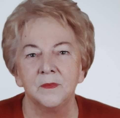 Portret starszej kobiety z krótkimi, jasnymi włosami, w czerwonej bluzce, z delikatnym uśmiechem i jasnym tłem.