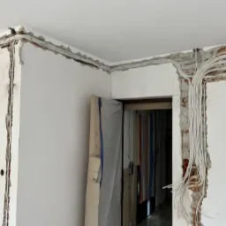 Instalacja elektryczna w bruzdach ściennych podczas remontu; widoczne kable w białych rurkach prowadzone wzdłuż ściany i pod sufitem, ściany częściowo otynkowane, fragment zakrytych drzwi.