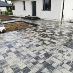 Ułożona, szaro-biała kostka brukowa przed domem w Wyszkowie. Widoczny fragment budynku z czarnymi oknami i drzwiami. Na palecie kostka do dalszych prac.