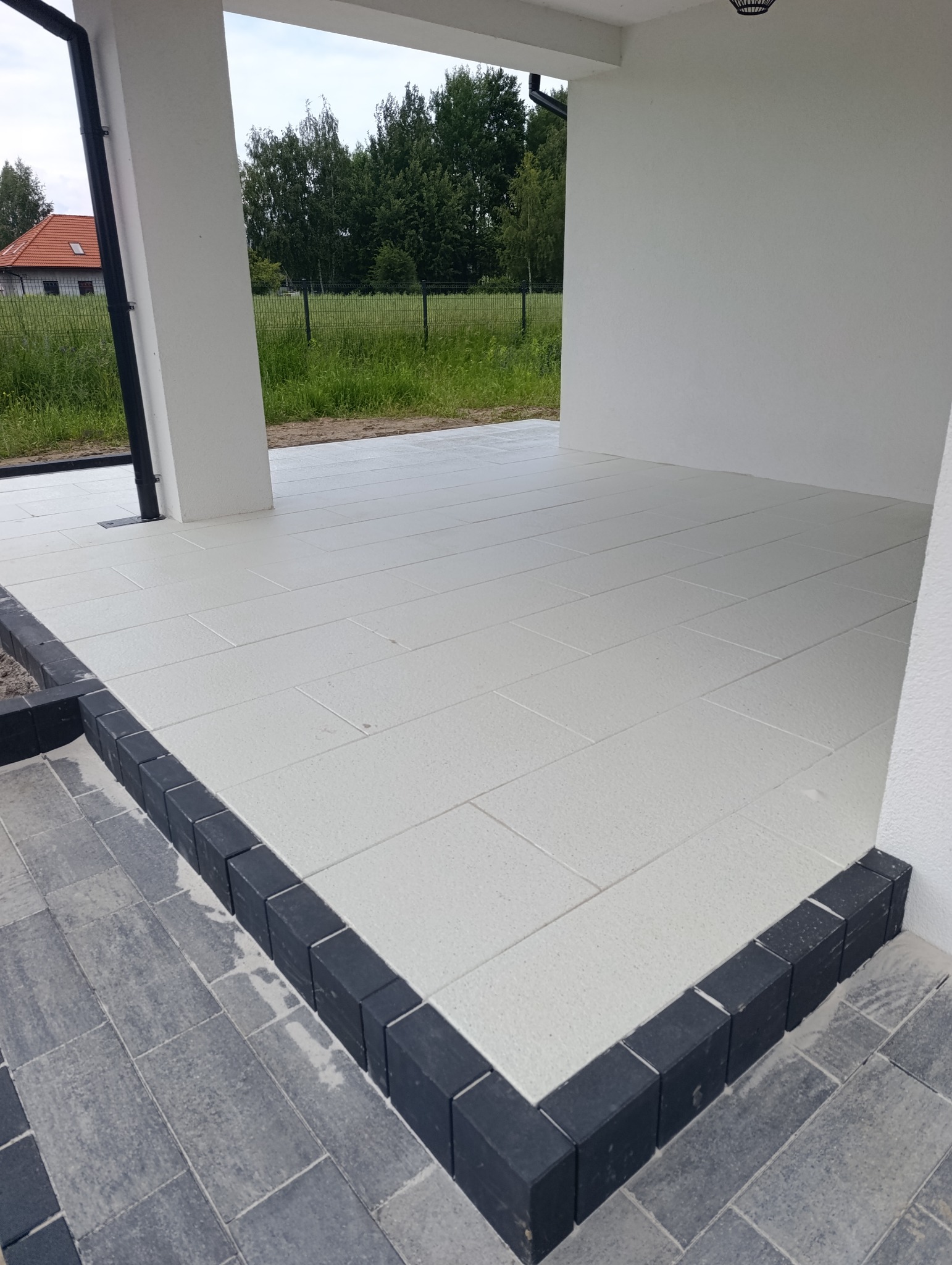 Biała, prostokątna kostka na tarasie domu, obramowana czarną kostką. Widok na ogród przez filary tarasu. Nowoczesne wykończenie.
