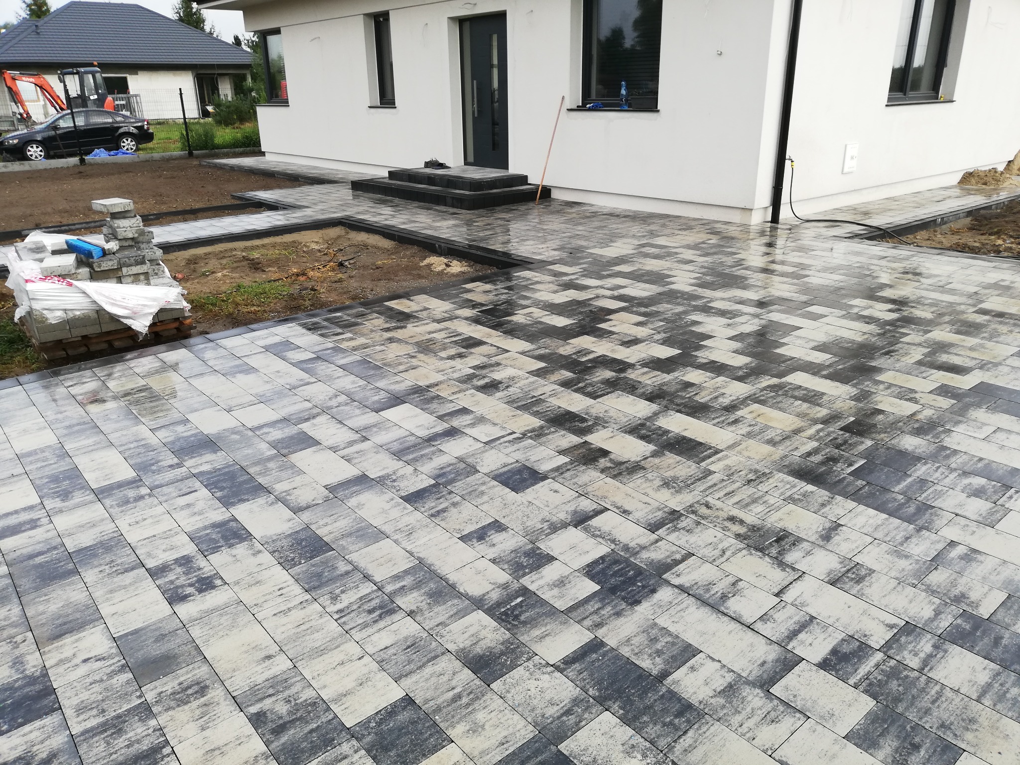 Ułożona, szaro-biała kostka brukowa przed domem w Wyszkowie. Widoczny fragment budynku z czarnymi oknami i drzwiami. Na palecie kostka do dalszych prac.