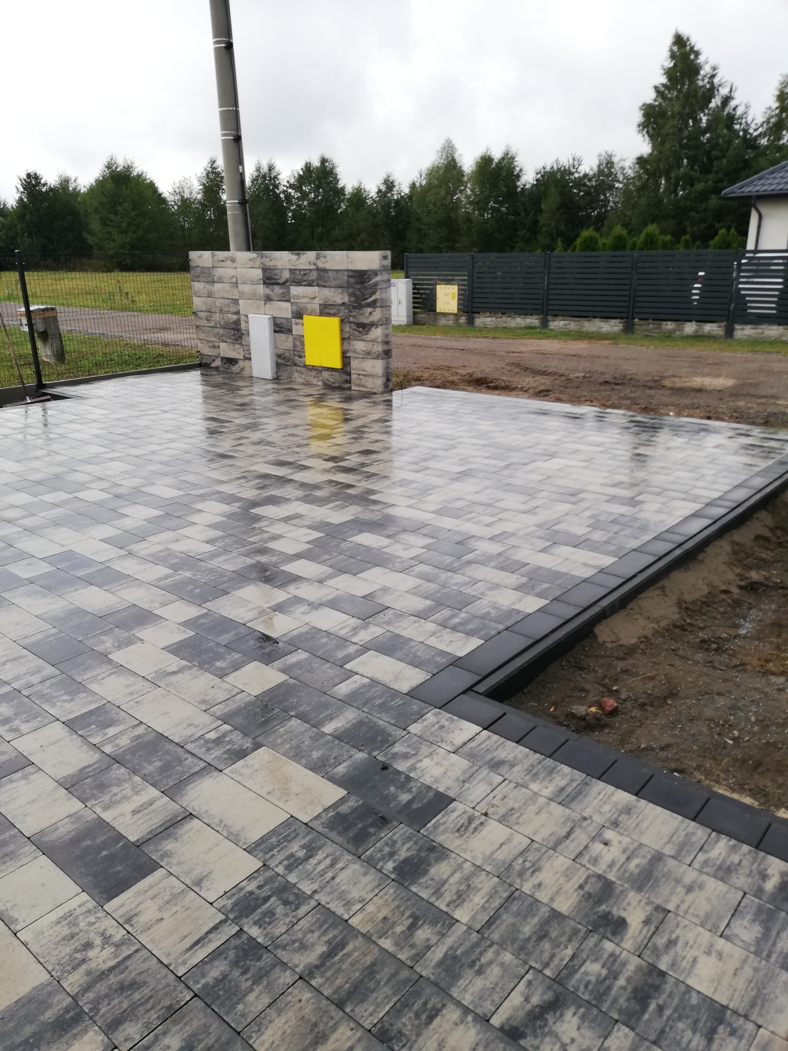 Ułożona kostka brukowa w odcieniach szarości i beżu, wzór szachownicy, obrzeża z ciemnej kostki. W tle słup elektryczny i dekoracyjna ściana z żółtym elementem.