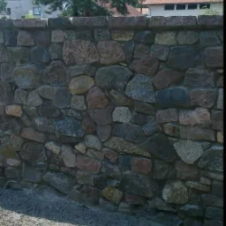 Murek ogrodzeniowy wykonany z nieregularnych kamieni polnych w odcieniach szarości i brązu, zwieńczony równą warstwą płaskich kamieni, z widocznym drzewem iglastym i budynkiem w tle.