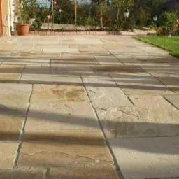 Jasna, kamienna nawierzchnia tarasu z widocznymi cieniami drzew, przylegająca do budynku z elementami kamiennej elewacji i ogrodem w tle.