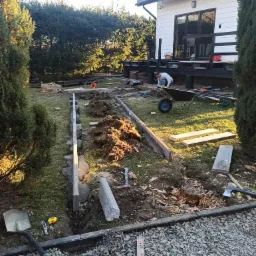 Przygotowanie terenu pod obrzeża trawnikowe z betonowych elementów, widoczny wykop, ułożone krawężniki, ziemia, narzędzia i pracownik z taczką w tle.