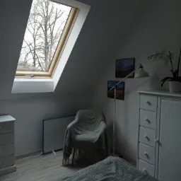 Pokój na poddaszu z oknem dachowym, przez które widać nagie gałęzie drzew; białe meble, krzesło przykryte pledem i lampa stojąca.