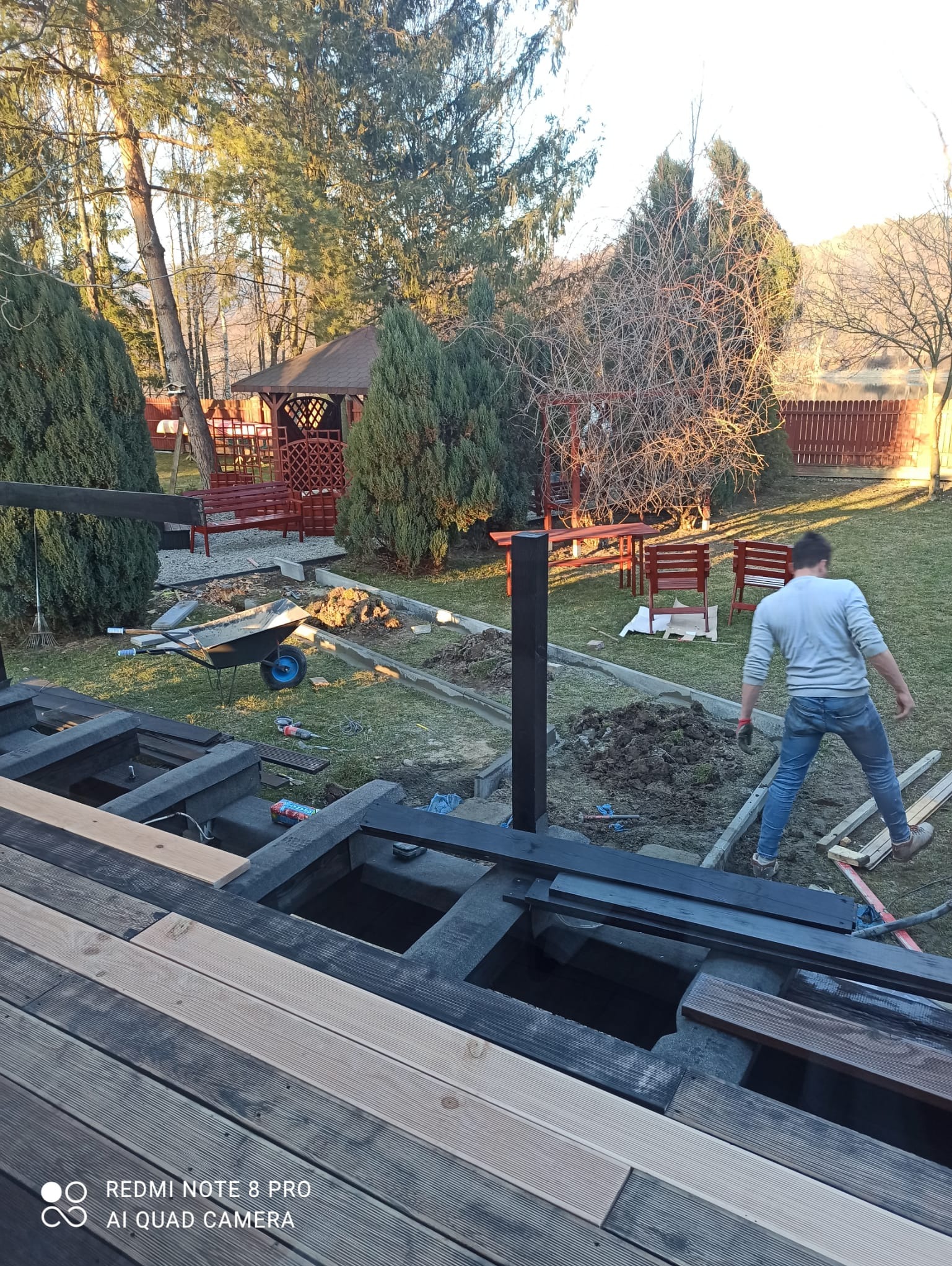 Budowa drewnianego tarasu na fundamencie betonowym w ogrodzie, widoczny pracownik w jeansach i wózek z narzędziami, w tle altana i drzewa.