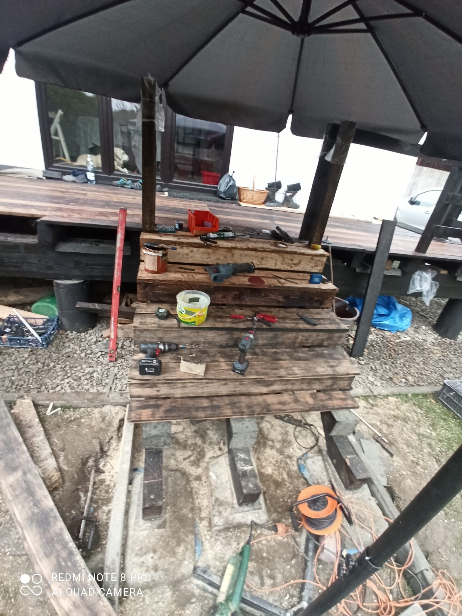 Budowa drewnianego tarasu: widoczne narzędzia, deski i elementy konstrukcyjne w trakcie montażu pod parasolem ogrodowym.