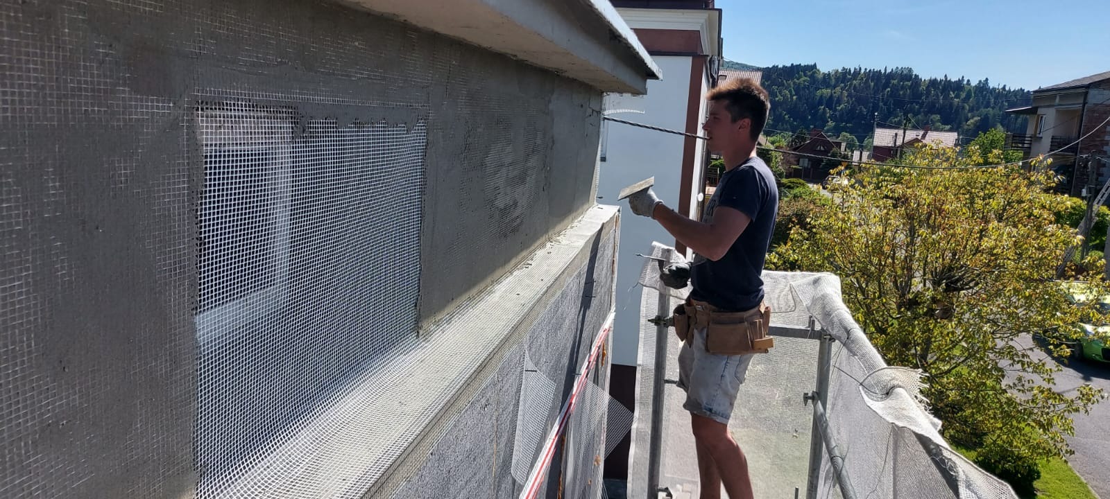 Pracownik w trakcie nakładania zaprawy na siatkę elewacyjną przy oknie, widok z rusztowania na tle zieleni.