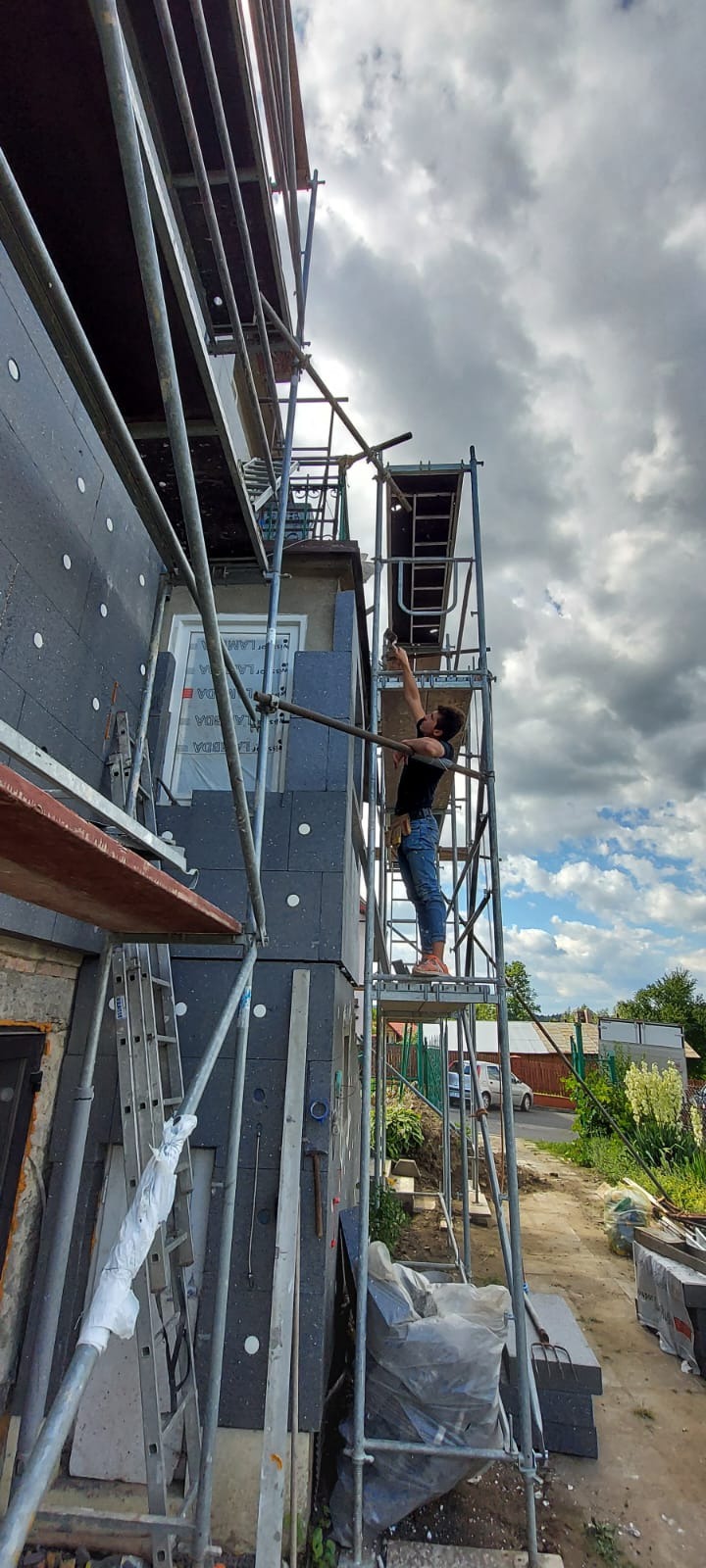 Pracownik na rusztowaniu, w trakcie montażu szarych płyt izolacyjnych na elewacji budynku, widoczne narzędzia i materiały budowlane, błękitne niebo z chmurami w tle.