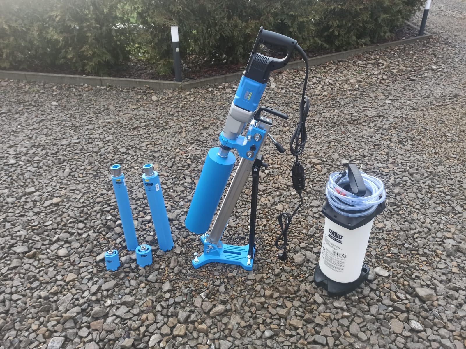Niebieska wiertnica rdzeniowa z zestawem wierteł i zbiornikiem na wodę, ustawiona na żwirowym podłożu przed zielonymi krzewami.