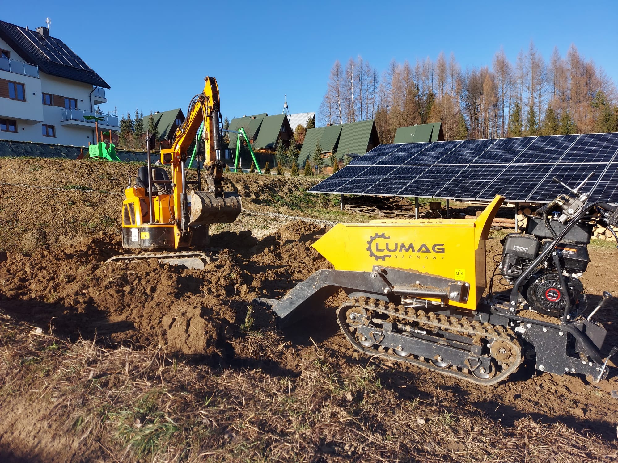 Żółta minikoparka i minitraktor gąsienicowy marki Lumag na placu budowy z widokiem na dom, domki letniskowe i panele słoneczne w słoneczny dzień.