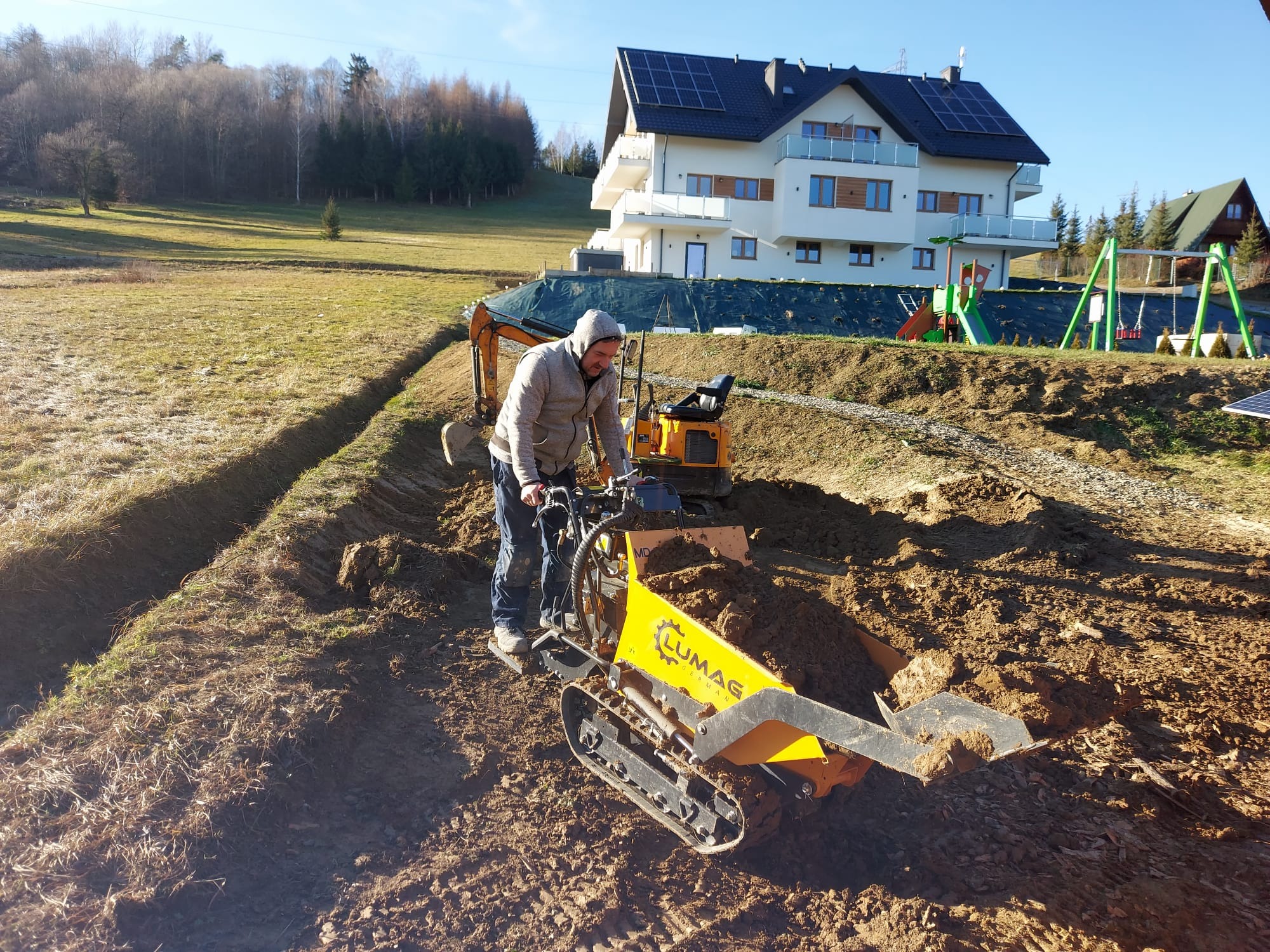 Operator obsługuje mini-wywrotkę gąsienicową marki Lumag podczas prac ziemnych na działce z nowym domem z panelami słonecznymi w tle.