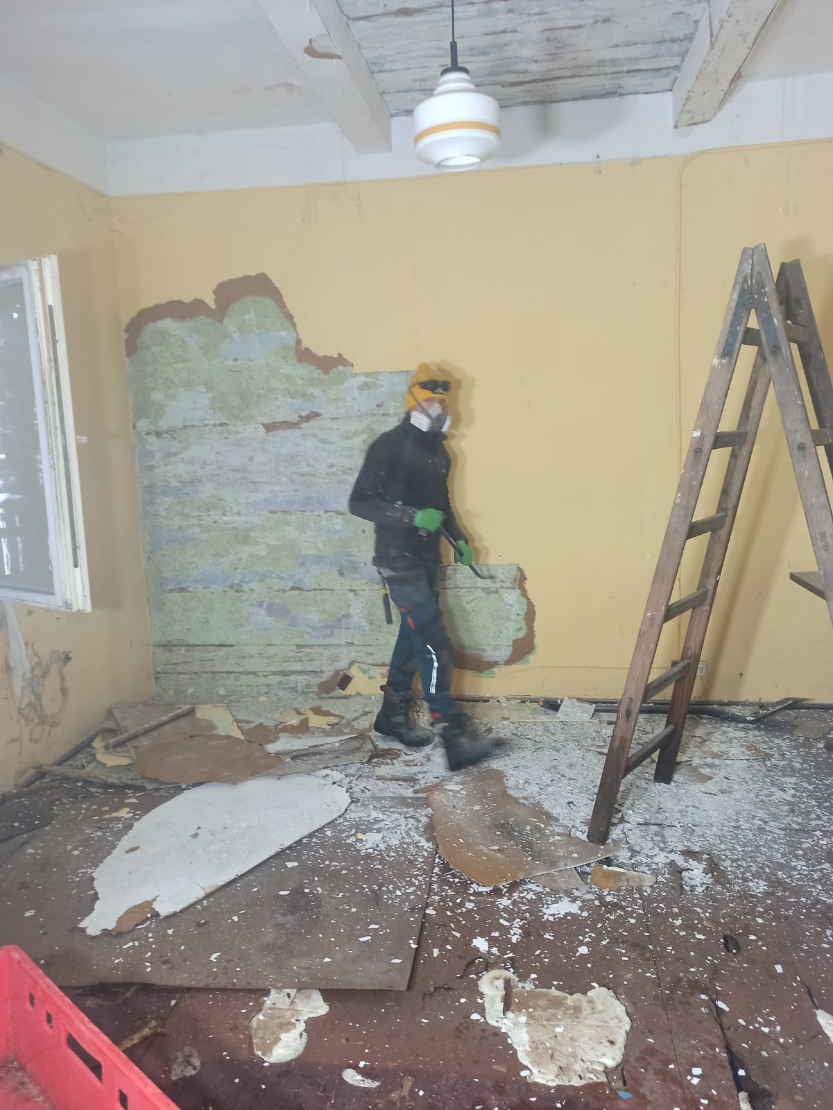 Pracownik w masce i okularach ochronnych usuwa starą warstwę ściany w pomieszczeniu z drewnianą drabiną i rozrzuconymi fragmentami gruzu na podłodze.