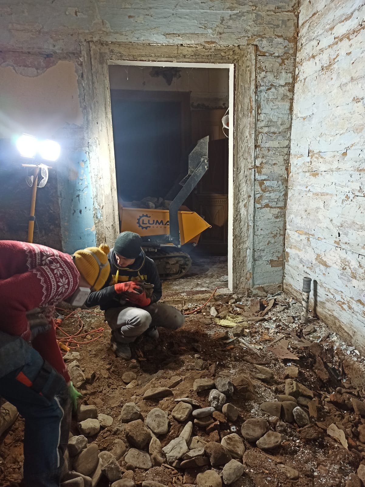 Dwóch pracowników w trakcie prac ziemnych wewnątrz starego budynku, widoczny minidumper Luma, oświetlenie halogenowe, odsłonięte kamienne fundamenty i fragmenty starej, łuszczącej się farby...