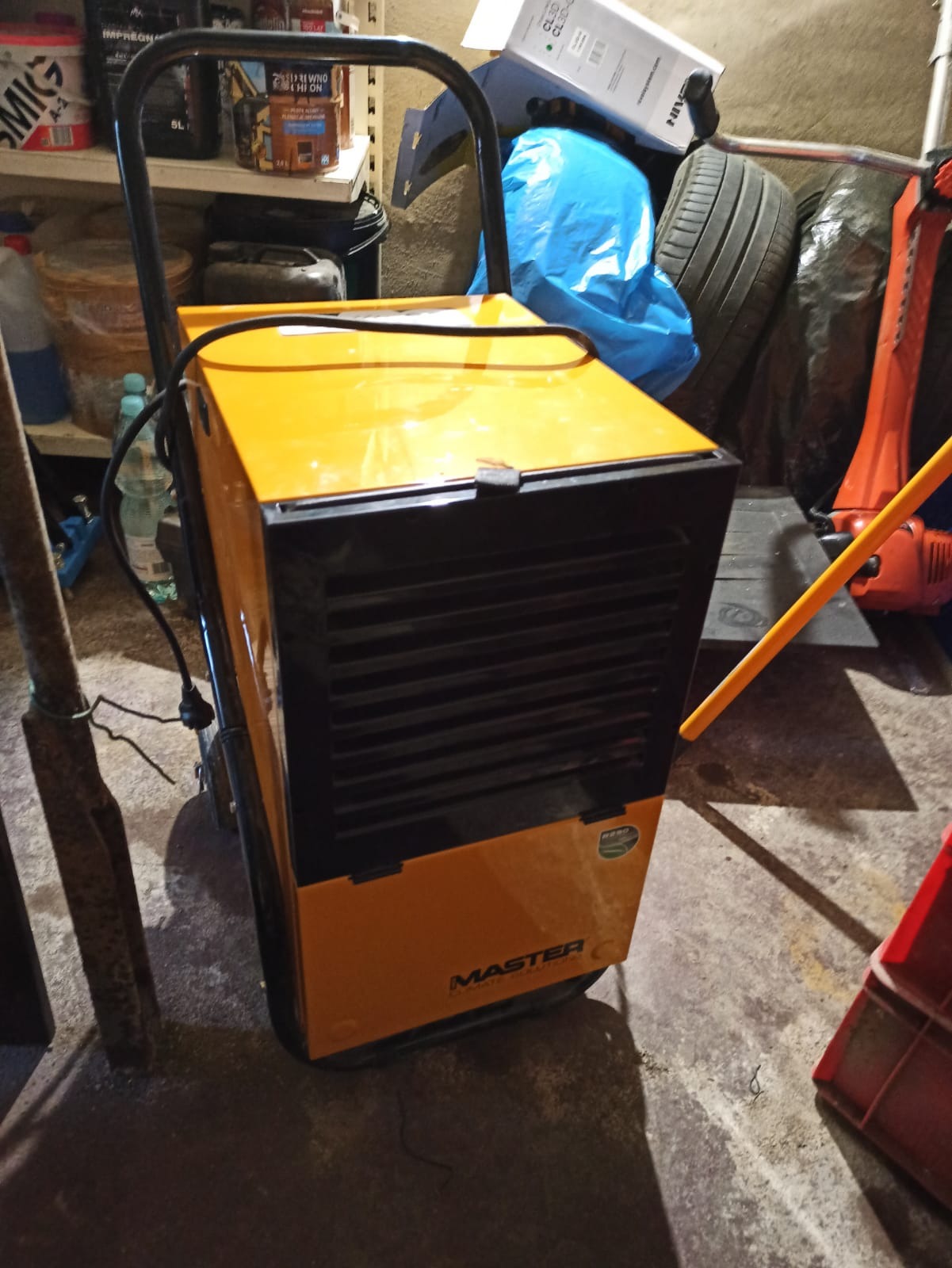 Pomarańczowo-czarny osuszacz powietrza marki Master na kółkach, stoi w zagraconym pomieszczeniu, widoczne narzędzia i inne przedmioty w tle.