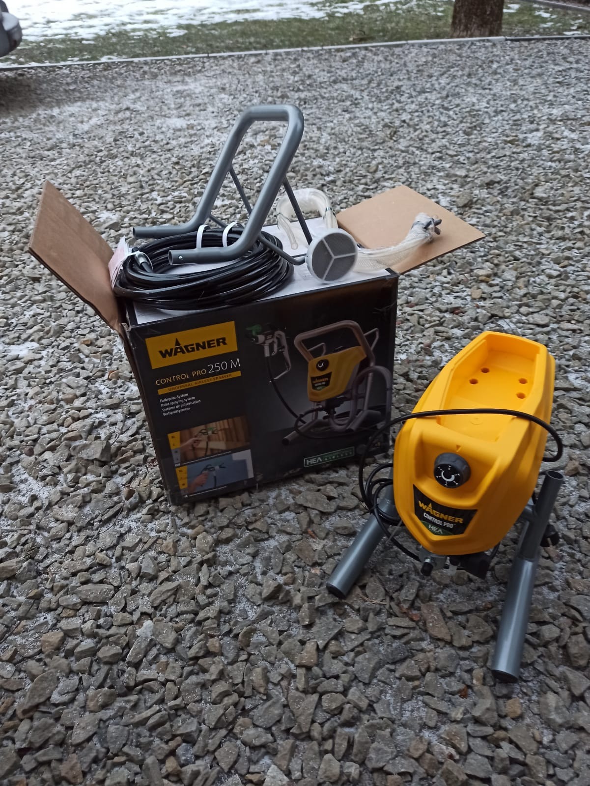 Rozpakowany, żółto-szary agregat malarski Wagner Control Pro 250 M na tle szarego żwiru, z widoczną czarną skrzynką z grafiką urządzenia i akcesoriami w kartonowym pudle.