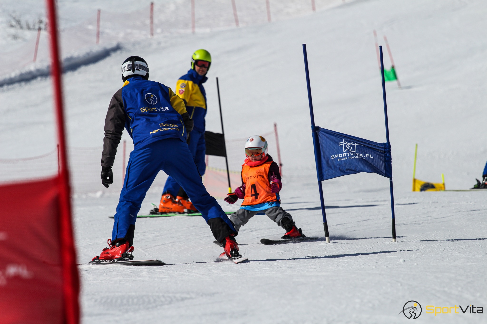 Instruktor narciarstwa uczy dziecko jazdy na nartach slalomem między tyczkami; flaga z logo SportVita.