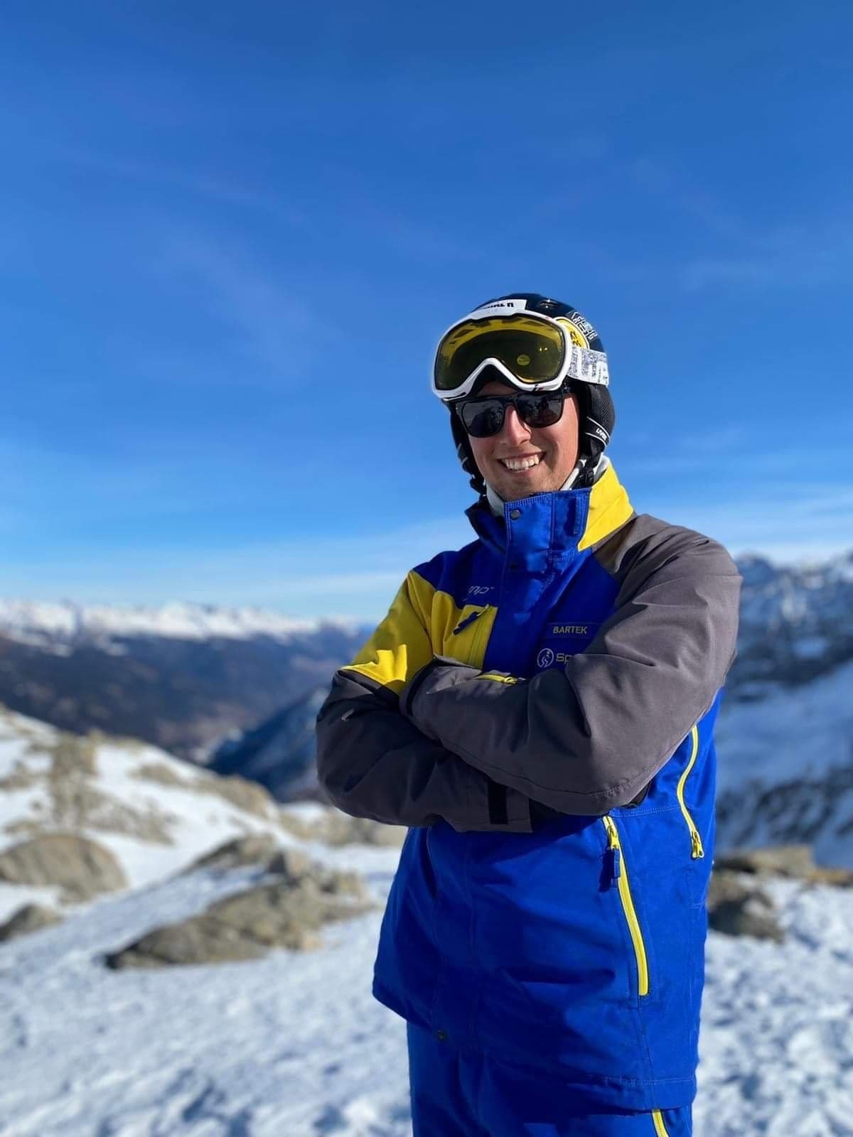 Uśmiechnięty mężczyzna w okularach przeciwsłonecznych i kasku narciarskim, ubrany w niebiesko-żółto-szarą kurtkę narciarską, stoi na szczycie góry z widokiem na ośnieżone szczyty w słoneczny dzień.