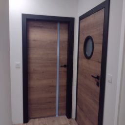 A.W.REM - Dwa drewnopodobne drzwi wewnętrzne z czarnymi ościeżnicami, jeden z okrągłym okienkiem, drugi z pionowym, niebieskim paskiem dekoracyjnym.