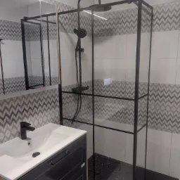 Nowoczesna łazienka z kabiną prysznicową typu walk-in z czarnymi ramami, umywalką nablatową na szafce z szufladami i płytkami w zygzakowaty wzór na ścianie.