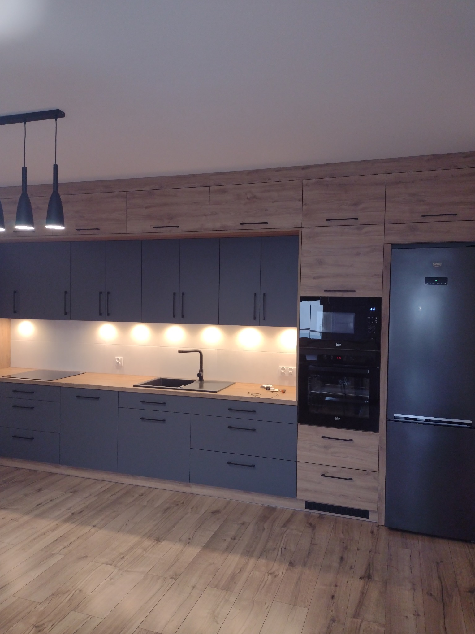 Nowoczesna kuchnia z szarymi szafkami dolnymi i drewnopodobnymi górnymi, czarnym zlewem i sprzętem AGD marki Beko, oświetlona punktowymi lampami podszafkowymi, z podłogą imitującą drewno.