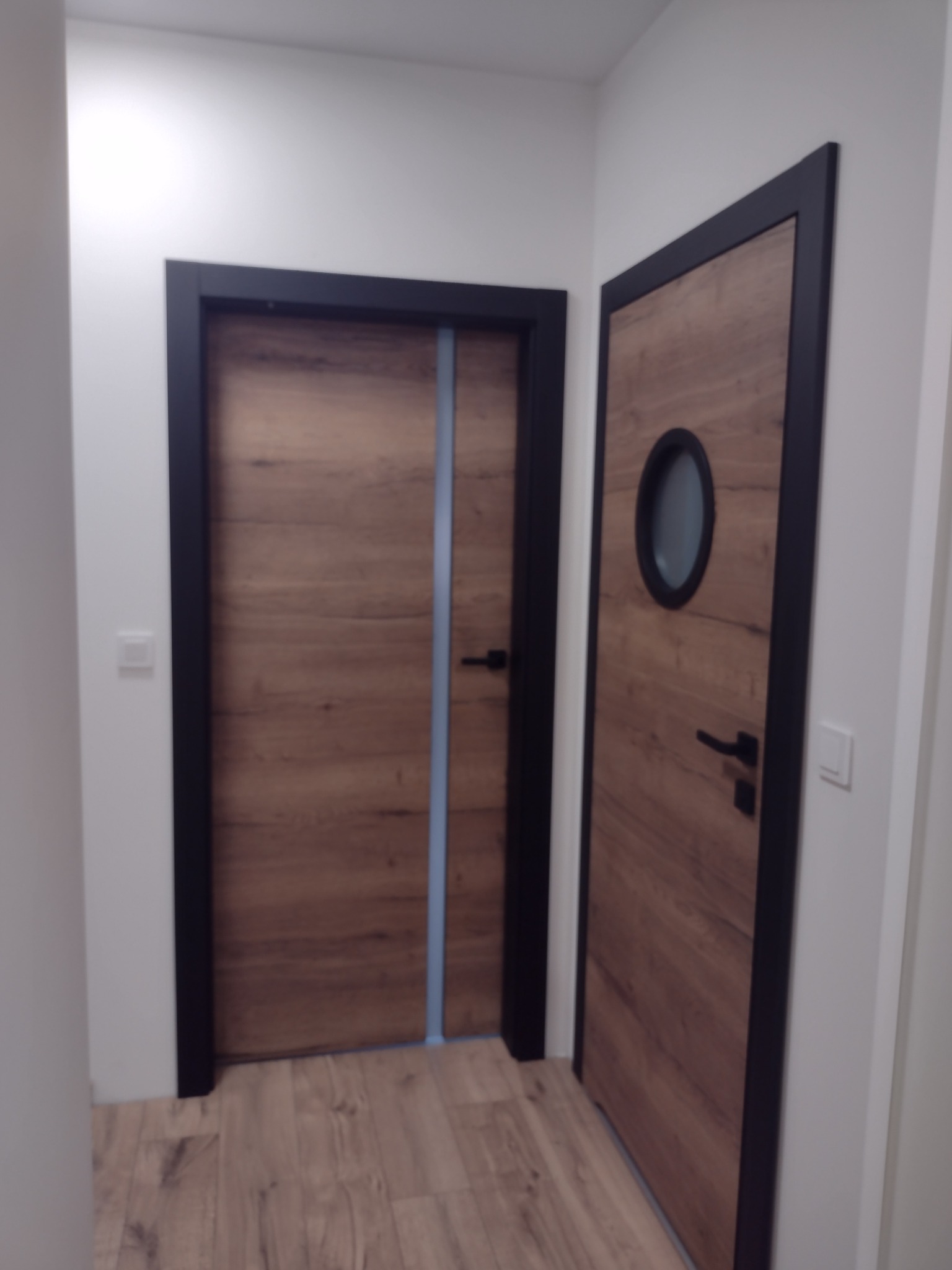 Dwa drewnopodobne drzwi wewnętrzne z czarnymi ościeżnicami, jeden z okrągłym okienkiem, drugi z pionowym, niebieskim paskiem dekoracyjnym.