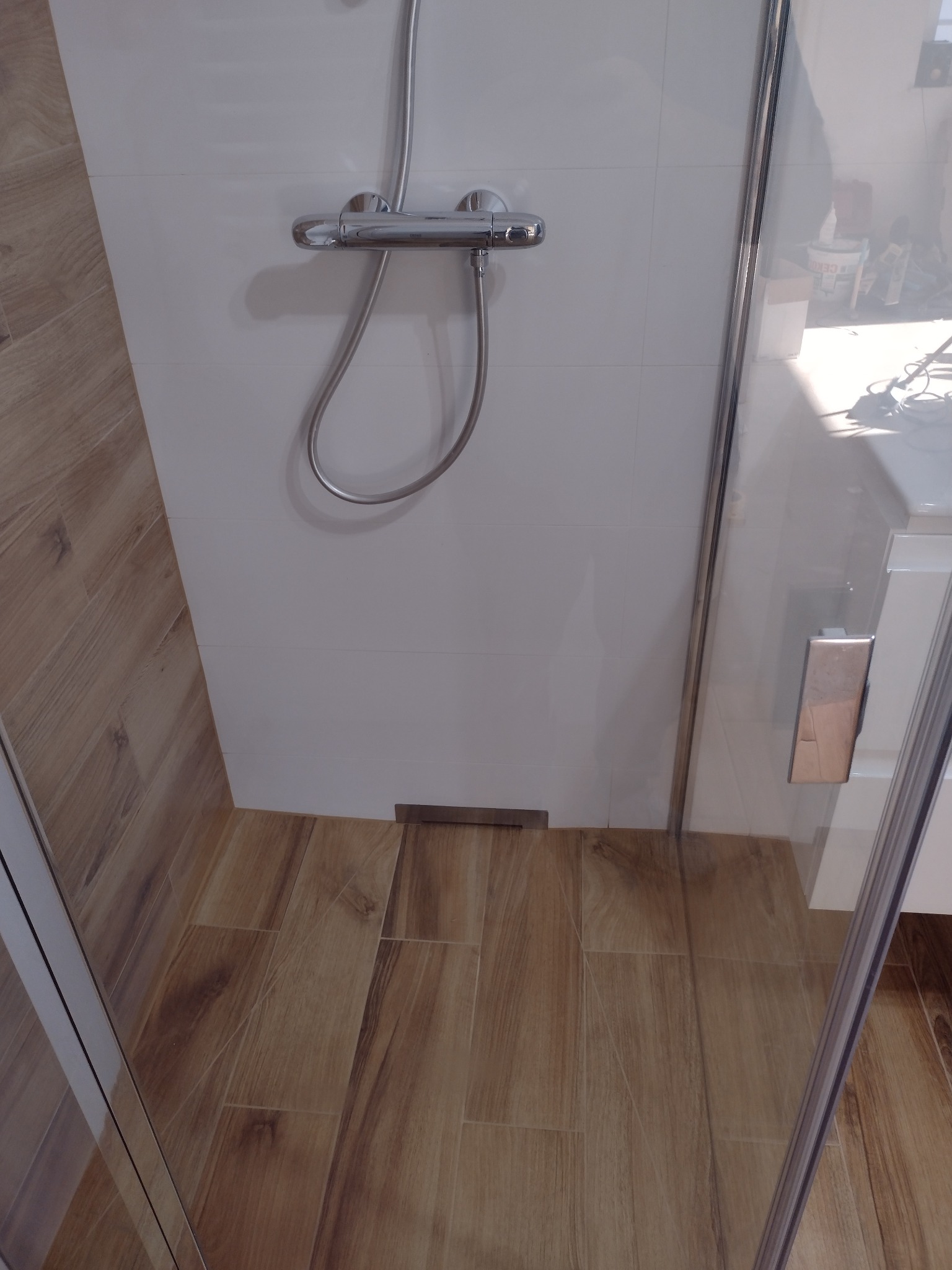 Kabina prysznicowa z chromowaną armaturą i odpływem liniowym, wykończona płytkami imitującymi drewno na podłodze i białymi płytkami na ścianach.