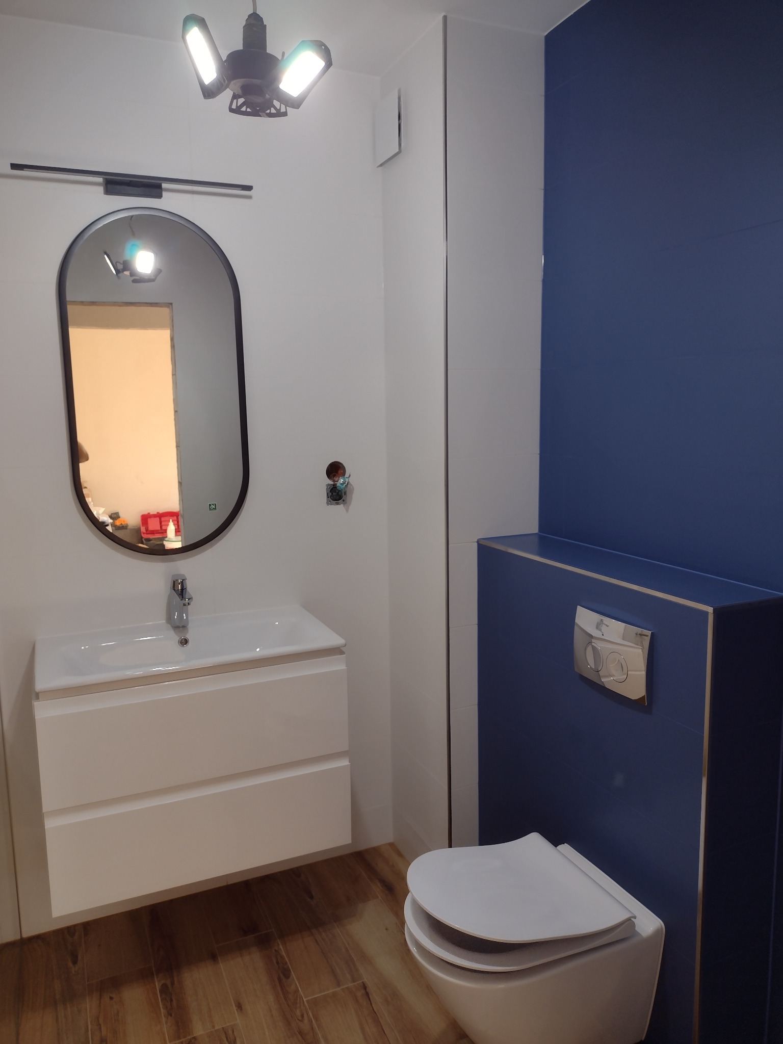 Nowoczesna łazienka z owalnym lustrem podświetlonym lampą, białą szafką z umywalką, podwieszaną toaletą na tle niebieskich płytek, i drewnopodobną podłogą.