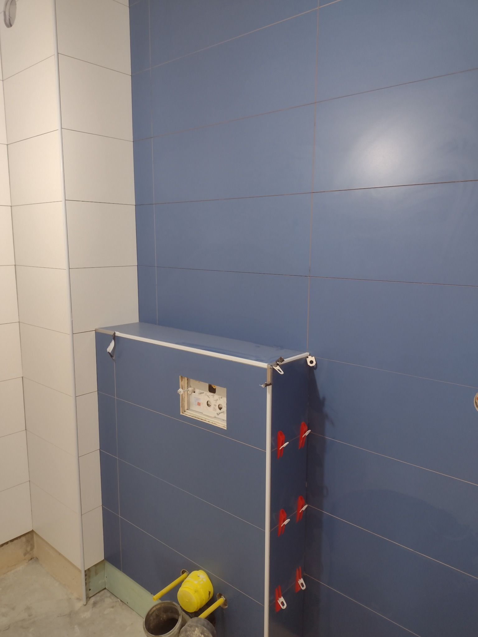 Niebiesko-biała łazienka w trakcie układania płytek, widoczne systemy poziomowania płytek, stelaż podtynkowy WC, żółty gumowy młotek i wiadro na podłodze.