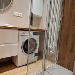 SIEMET REMONTY & WYKOŃCZENIA. - Nowoczesna łazienka z kabiną prysznicową ze szkła hartowanego, białą pralką Electrolux i grzejnikiem drabinkowym na ścianie z drewnopodobnymi panelami.