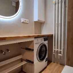 Nowoczesna łazienka z drewnopodobną podłogą, okrągłym lustrem z podświetleniem LED, pralką Electrolux zabudowaną pod drewnianym blatem i białym grzejnikiem drabinkowym.