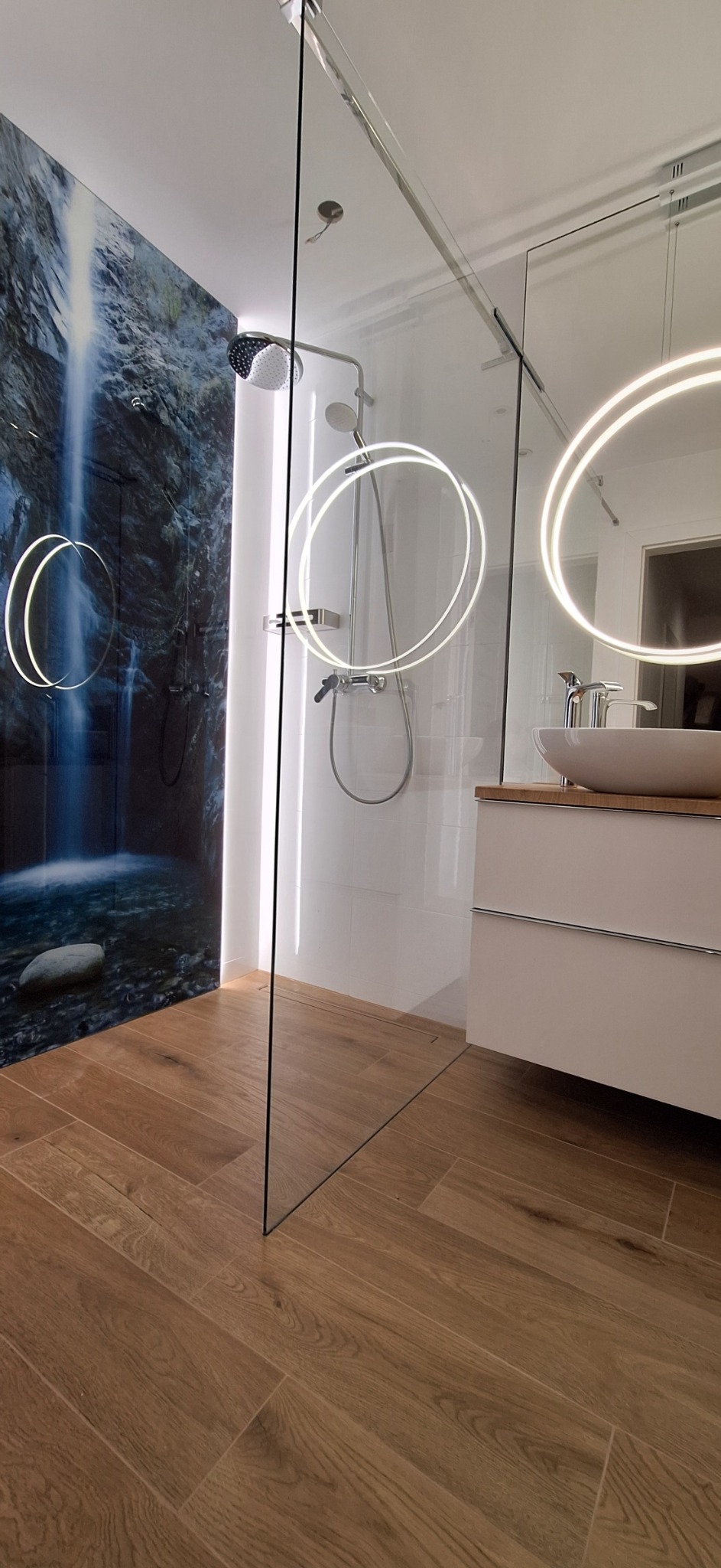Nowoczesna łazienka z kabiną prysznicową typu walk-in, drewnopodobnymi płytkami na podłodze ułożonymi w jodełkę oraz panelem ściennym z motywem wodospadu.