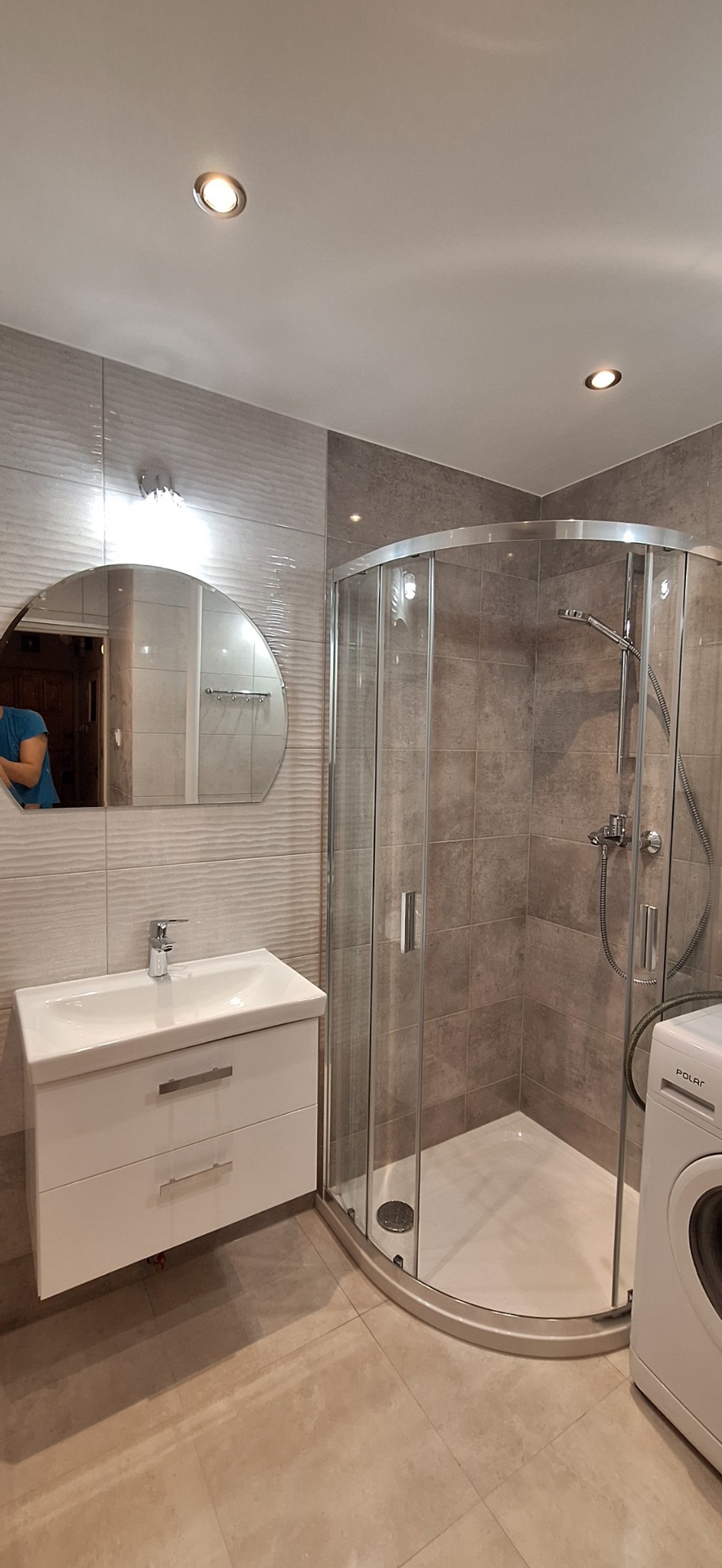 Nowoczesna łazienka z kabiną prysznicową narożną, umywalką z szafką w kolorze białym, półokrągłym lustrem i pralką marki Polar.