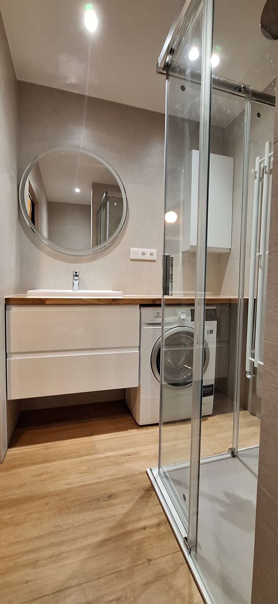 Nowoczesna łazienka w jasnych kolorach z owalnym lustrem, umywalką z szafką, pralką i kabiną prysznicową z przesuwnymi drzwiami.