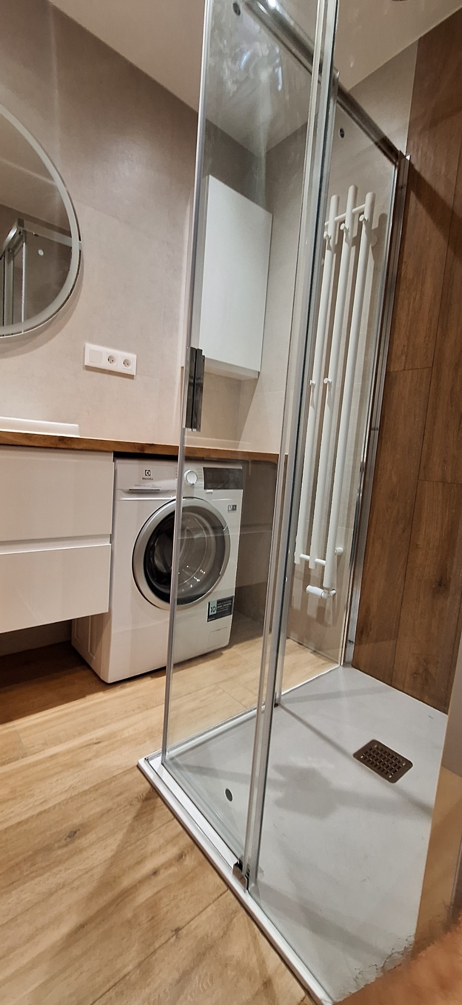 Nowoczesna łazienka z kabiną prysznicową ze szkła hartowanego, białą pralką Electrolux i grzejnikiem drabinkowym na ścianie z drewnopodobnymi panelami.